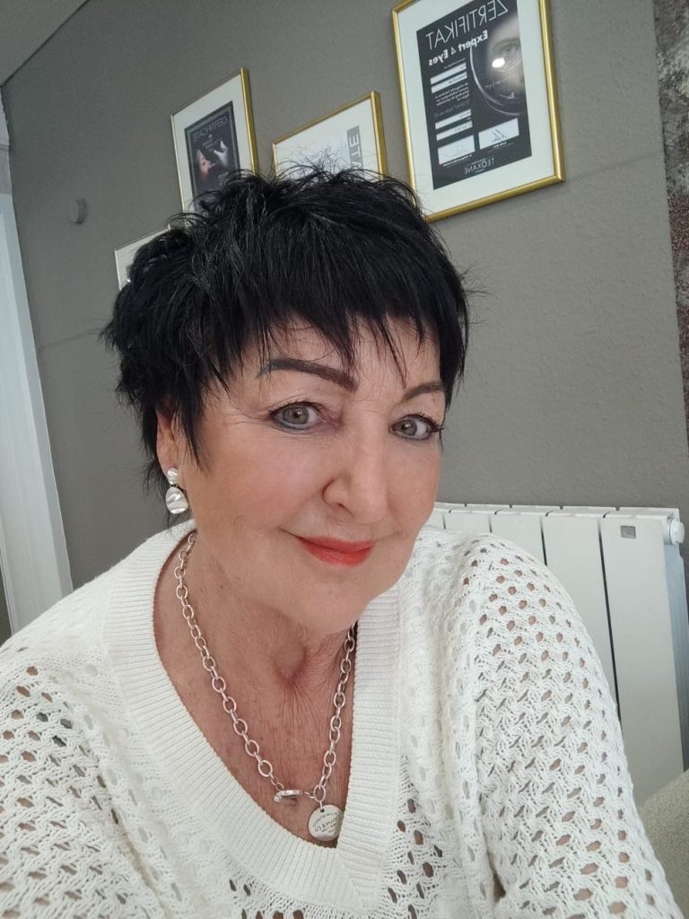 Porträt einer Frau mit kurzen dunklen Haaren, hellen Lippen und Schmuck, in einem hellen Raum.